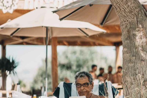 Guest comfortably seated under a tree, reading a newspaper at Naxaki Beach Lounge, with the restaurant's sign in the background