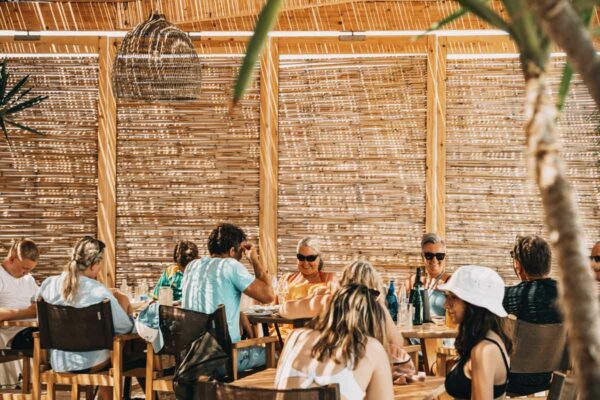 People enjoying a meal under a shaded bamboo structure at a beachside restaurant