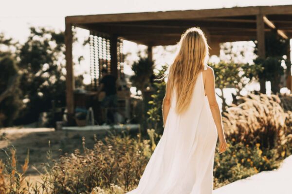 A woman in a flowing white dress walking through the garden area of Naxaki Beach Lounge on a sunny day