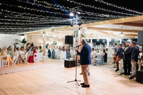 A wedding guest giving a speech under twinkling string lights at Naxaki Beach Lounge and Restaurant, with seated guests enjoying the reception